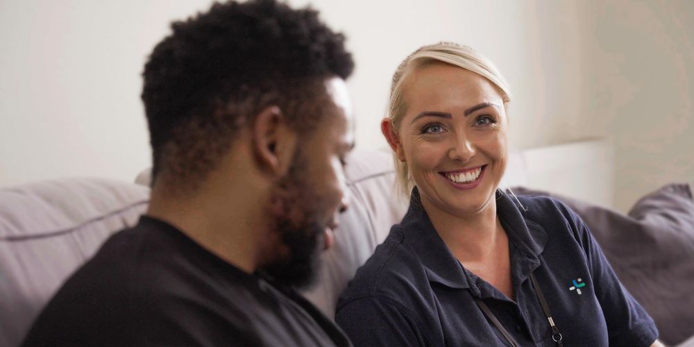 Man and woman sitting on a couch; the woman, in a dark shirt, is smiling warmly at the man.