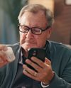 Elderly man on a couch examines a pill bottle while looking at his smartphone, wearing glasses and a sweater.