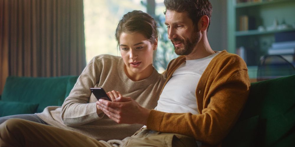 A couple sitting on a couch, looking at a smartphone together, with a bright window and shelves in the background.