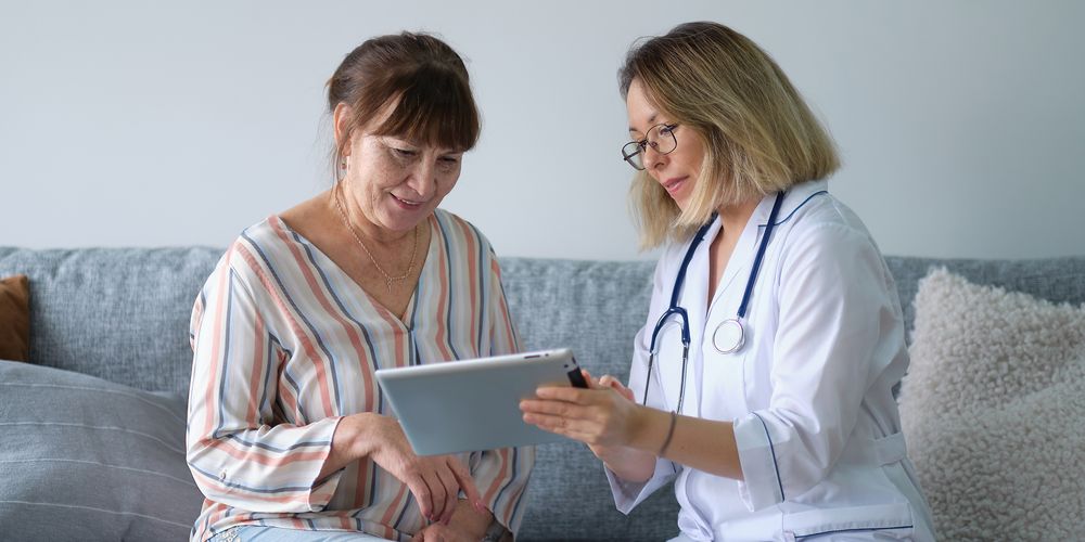 Doctor showing a tablet to a patient on a couch, both engaged in discussion. The doctor wears a stethoscope and glasses.
