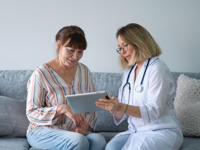 A doctor in a white coat shows a tablet to a woman seated on a sofa.
