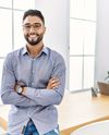 Smiling man with glasses and beard stands confidently in a bright office with folded arms, behind a wooden table, plant, and laptop.
