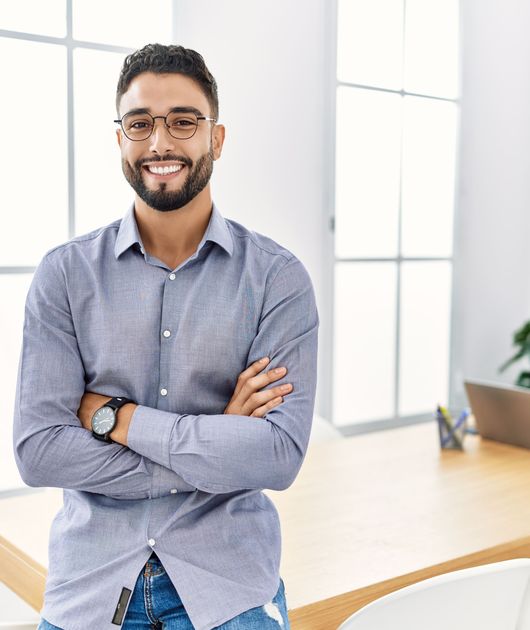 Smiling man with glasses and beard stands confidently in a bright office with folded arms, behind a wooden table, plant, and laptop.