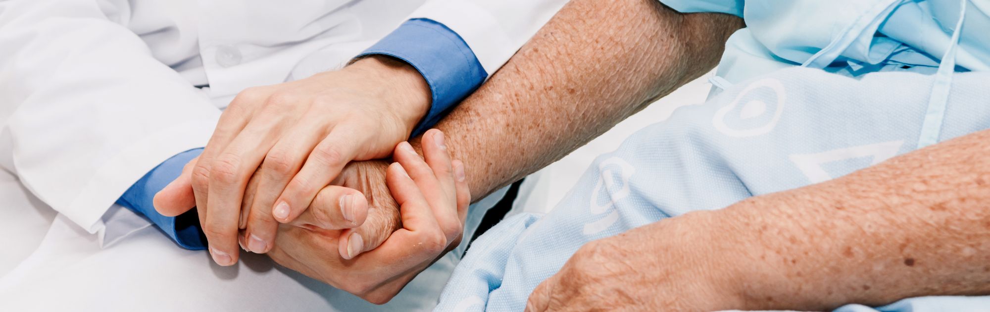 A doctor in a white coat holds hands with a patient in a light blue hospital gown, offering comfort and support.