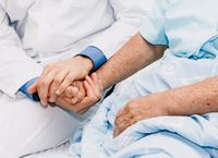 A doctor in a white coat holds hands with a patient in a light blue hospital gown, offering comfort and support.