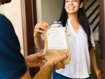 Smiling woman at door hands brown paper bag to a person.