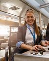 Smiling female airport staff at a check-in desk, using a computer, with colleagues working in the background.
