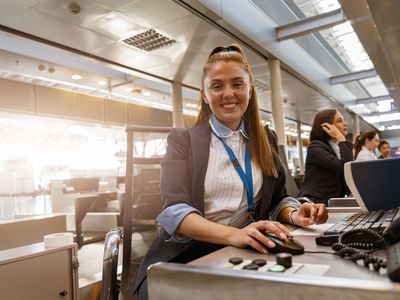 Smiling female airport staff at a check-in desk, using a computer, with colleagues working in the background.