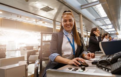 Smiling female airport staff at a check-in desk, using a computer, with colleagues working in the background.