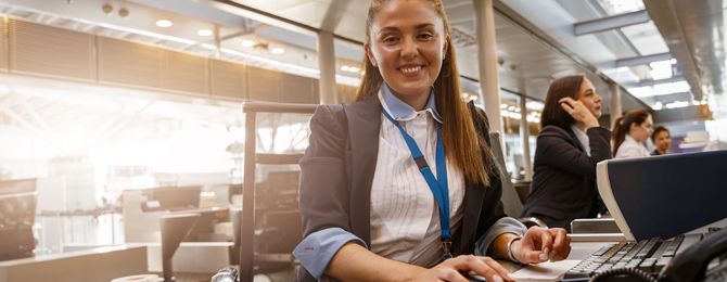 Smiling female airport staff at a check-in desk, using a computer, with colleagues working in the background.