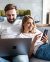 Couple sitting on a couch, smiling and using a laptop and smartphone.
