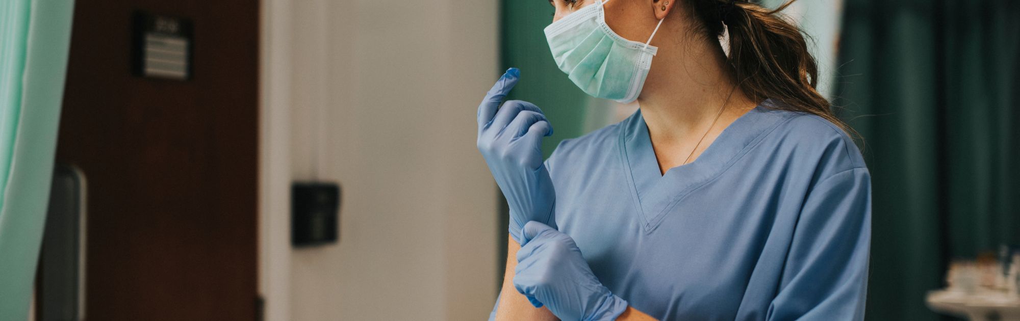Healthcare professional in blue scrubs and mask adjusts gloves.