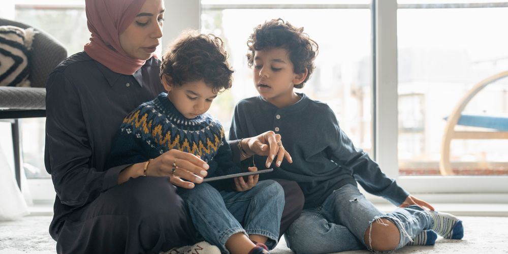 A woman in a hijab sits on the floor with two young children, looking at a tablet together in a cozy, sunlit room.