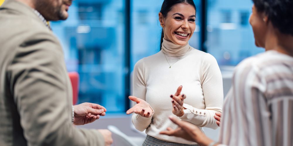 Three people engaged in a cheerful conversation in a modern office setting, with large windows in the background.