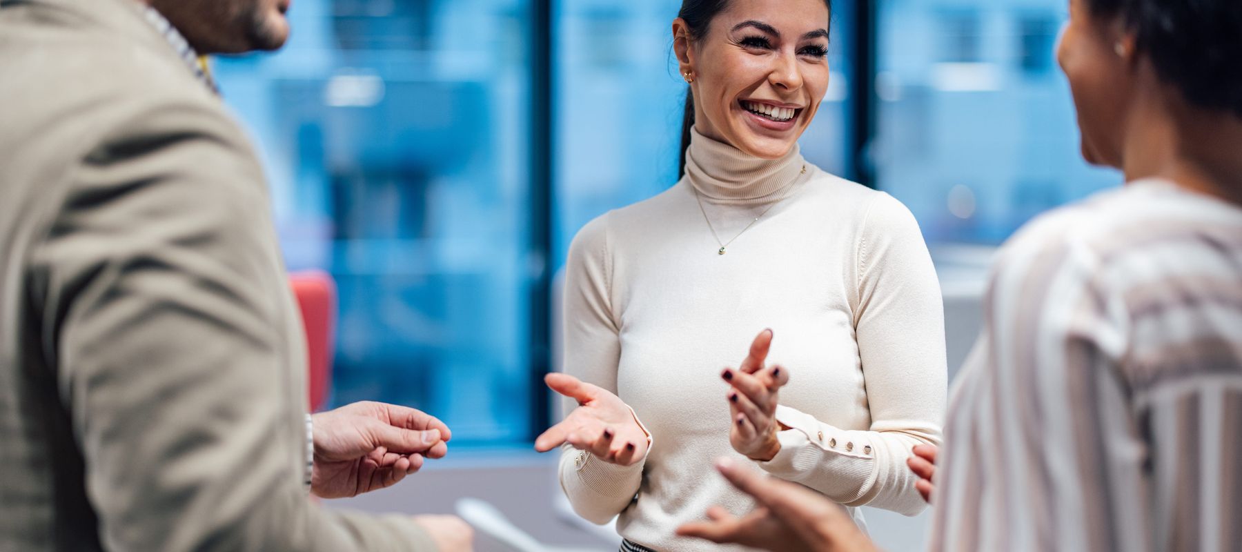 Three people engaged in a cheerful conversation in a modern office setting, with large windows in the background.
