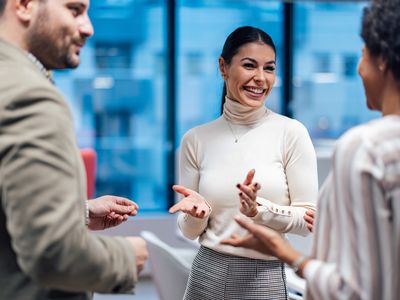 Three people engaged in a cheerful conversation in a modern office setting, with large windows in the background.