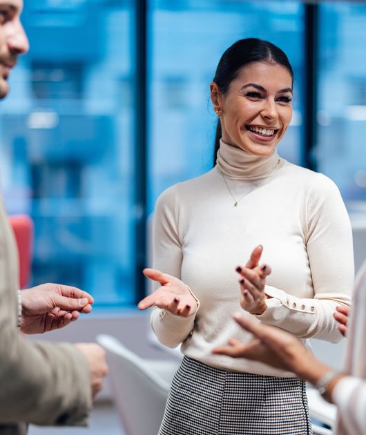 Three people engaged in a cheerful conversation in a modern office setting, with large windows in the background.