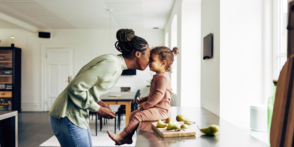 Woman kissing a child on the forehead as the child sits on a kitchen counter, surrounded by pears, in a bright and modern kitchen.