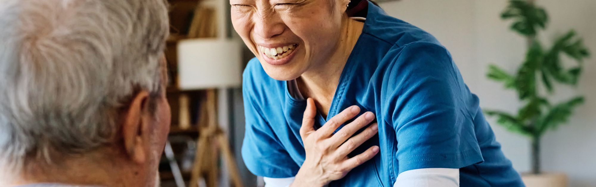 A nurse in blue scrubs smiles warmly, placing a hand on an elderly man's shoulder in a cozy living room.