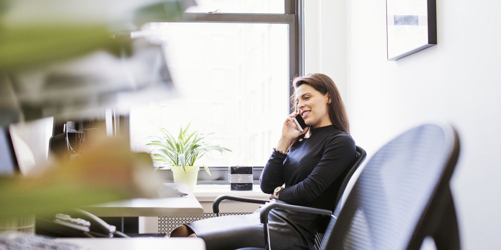 Woman in a black outfit sitting at a desk, talking on the phone, smiling, with a plant by the window in a bright office.
