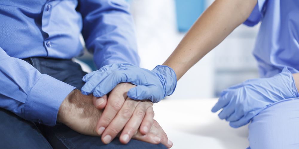 Close-up of a healthcare professional wearing gloves gently holding a patient’s hands, offering support in a clinical setting.