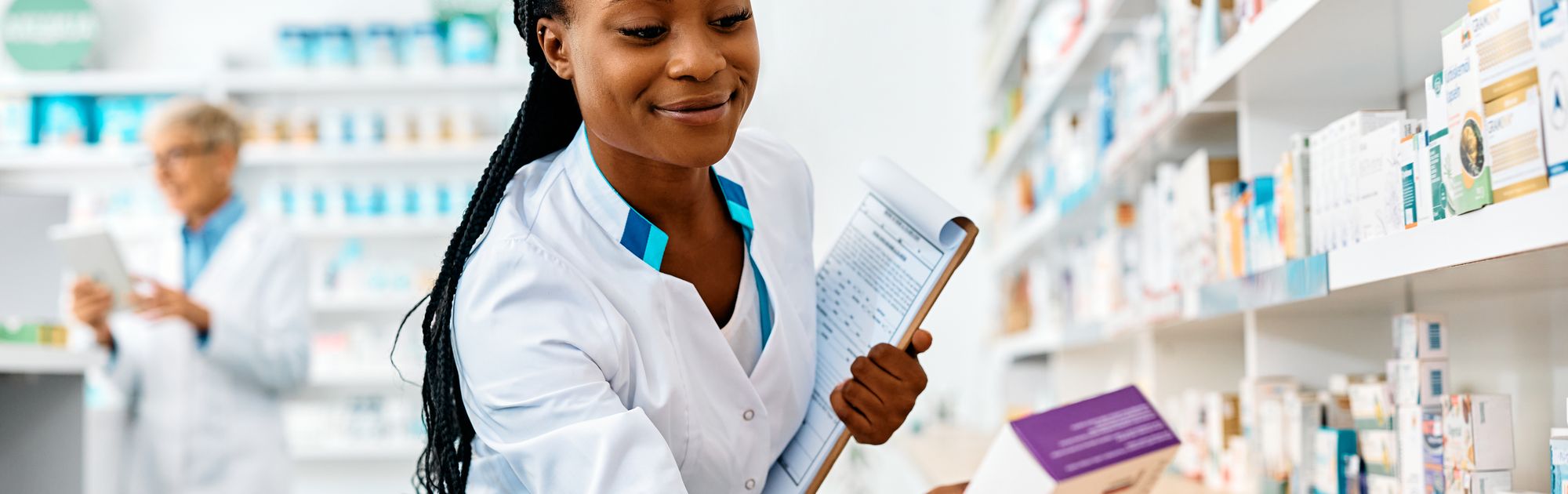 A pharmacist in a white coat selects a medication box from a shelf while holding a clipboard. Another pharmacist is in the background.