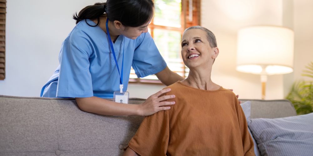 Healthcare professional in blue scrubs gently placing hands on a seated patient’s shoulders while speaking with them in a home living room setting.
