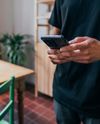 Person in a black shirt holding a smartphone in a cozy room with wooden furniture and a potted plant nearby.