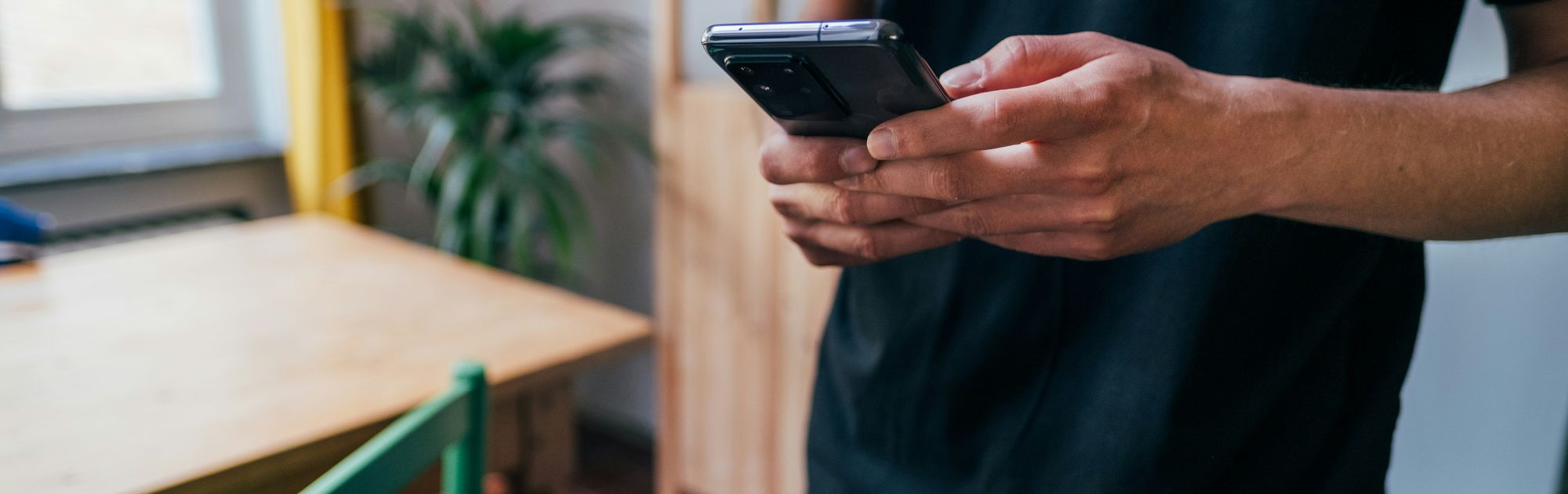 Person in a black shirt holding a smartphone in a cozy room with wooden furniture and a potted plant nearby.