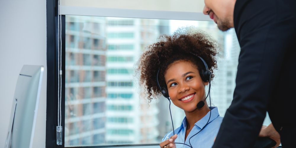 Woman wearing a headset smiles at a colleague in an office with a city view.