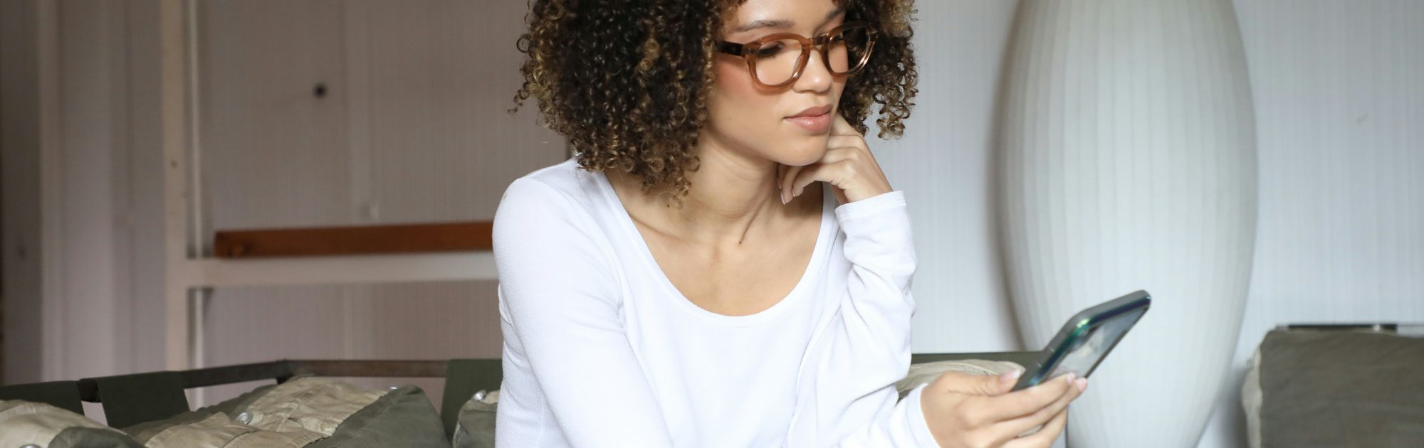 Person with curly hair and glasses looks at a phone while sitting on a sofa in a cozy room. They wear a white long-sleeve shirt and jeans.