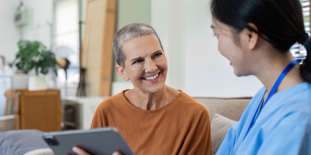 A nurse in blue scrubs chats happily with a smiling woman in an orange shirt, holding a tablet in a cozy living room setting.