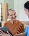 Woman in casual wear smiles at a healthcare worker holding a tablet, sitting together in a cozy room.