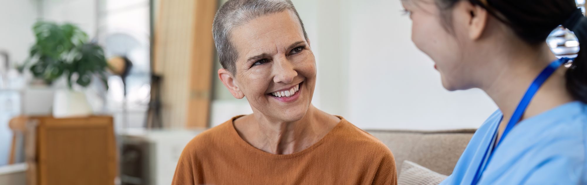 Woman in casual wear smiles at a healthcare worker holding a tablet, sitting together in a cozy room.