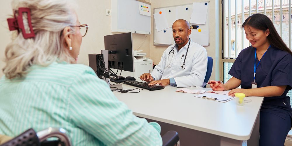 Doctor seated at a desk using a computer while consulting with a patient in a wheelchair, with a healthcare professional taking notes beside them in a clinical setting.