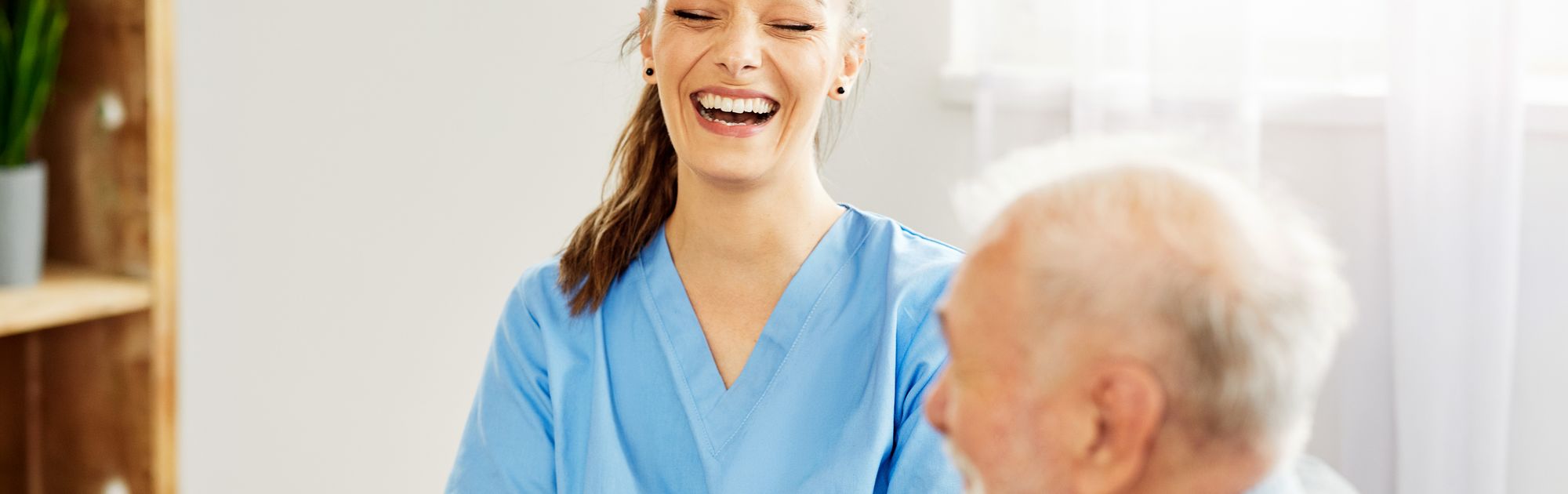 Healthcare professional in blue scrubs smiling while speaking to an older patient during a home care visit, with the patient seated in the foreground.