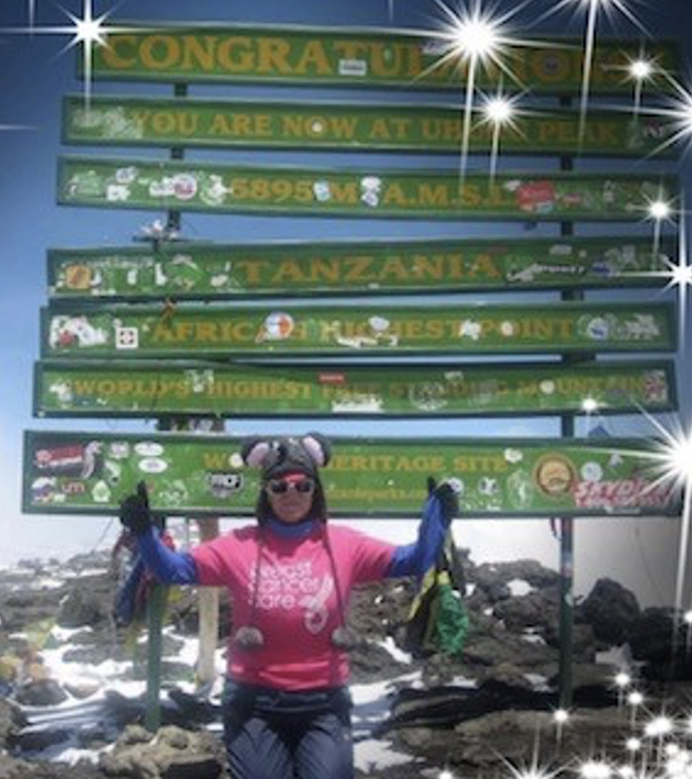Person wearing a pink shirt and animal hat at the Uhuru Peak sign on Mount Kilimanjaro, surrounded by sparkles.