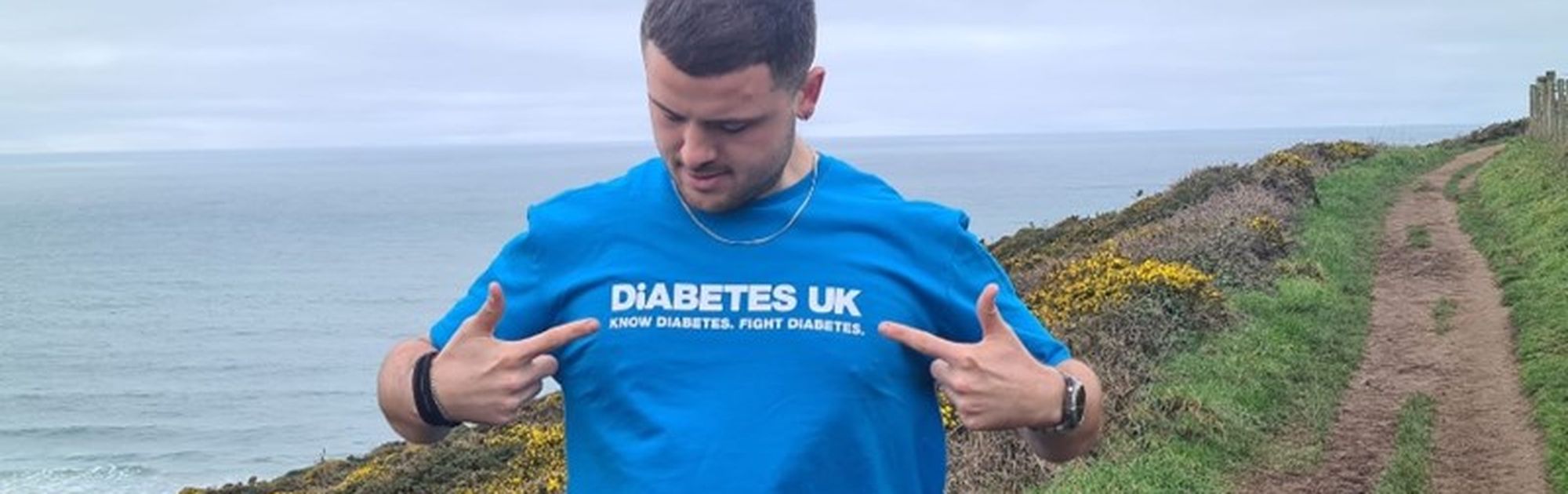 Man in a blue "Diabetes UK" shirt stands on a coastal path, pointing to the shirt. Ocean and cloudy sky in the background.