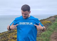 Man in a blue "Diabetes UK" shirt stands on a coastal path, pointing to the shirt. Ocean and cloudy sky in the background.