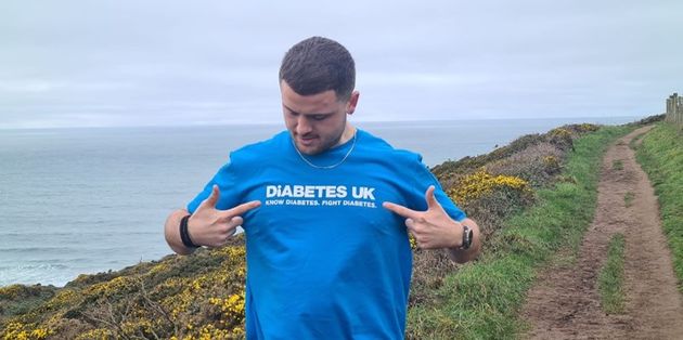 Man wearing a "Diabetes UK" shirt points at its logo, standing on a coastal path with the sea and cloudy sky in the background.