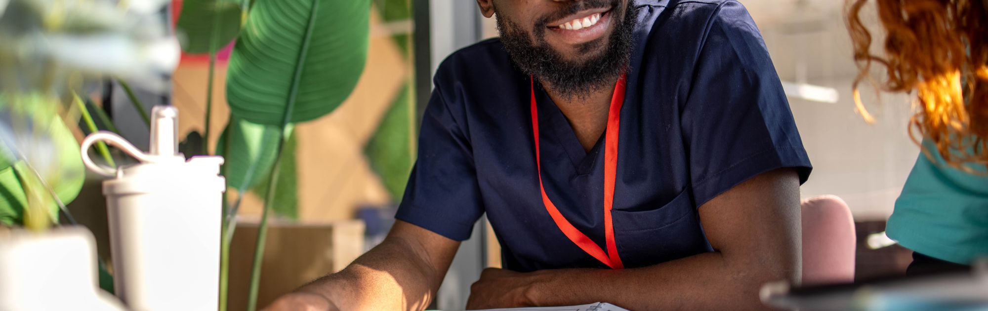 Man in navy scrubs smiles while sitting at a table with open notebooks and green plants in the background.