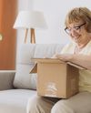 Elderly woman smiling as she opens a cardboard box on a couch, with a lamp and plant nearby.
