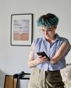 Person with teal curly hair, wearing a striped shirt, looks at a phone in a cozy, art-adorned room.