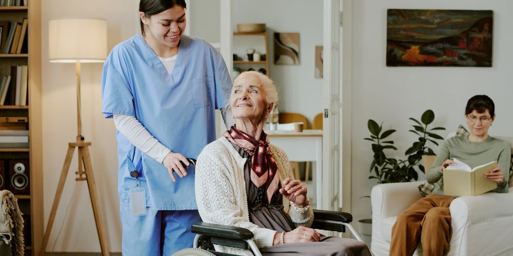 A caregiver assists an elderly woman in a wheelchair. Another person reads on a couch in a cozy, well-lit living room with plants and books.