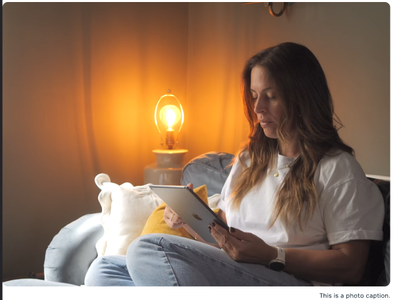 Woman sitting on a couch, reading a tablet beside a lit lamp.