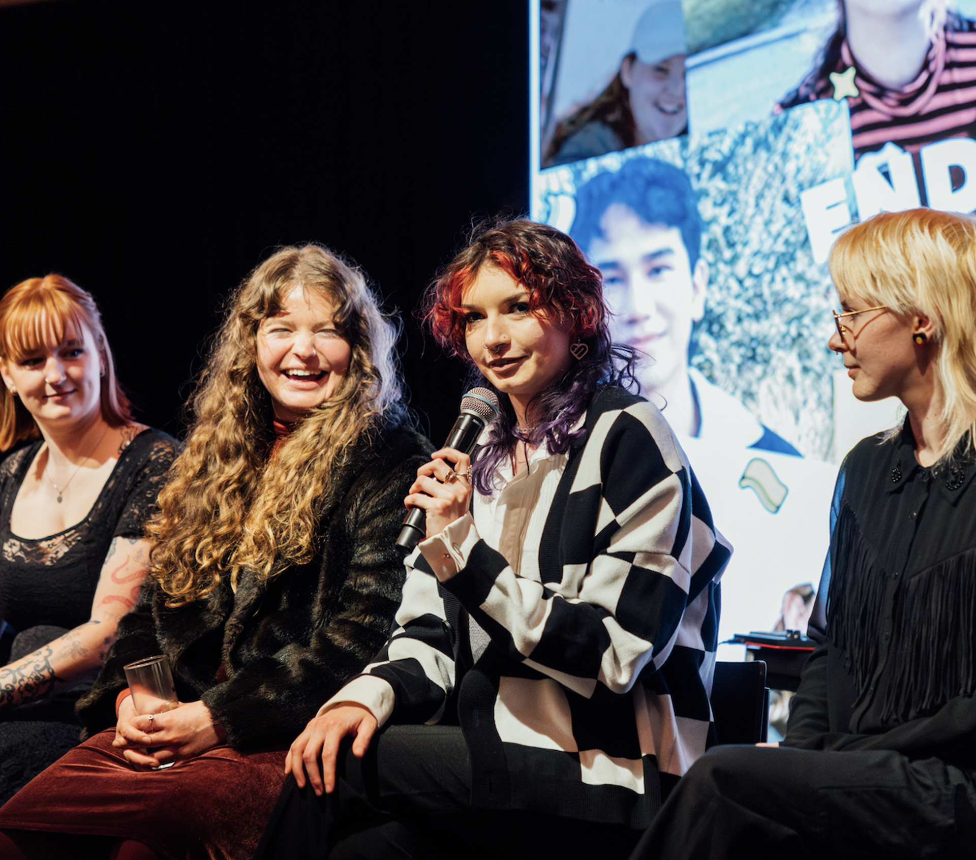 Four people seated on a panel discussion. One is speaking into a microphone, others are smiling. A large collage of photos is in the background.
