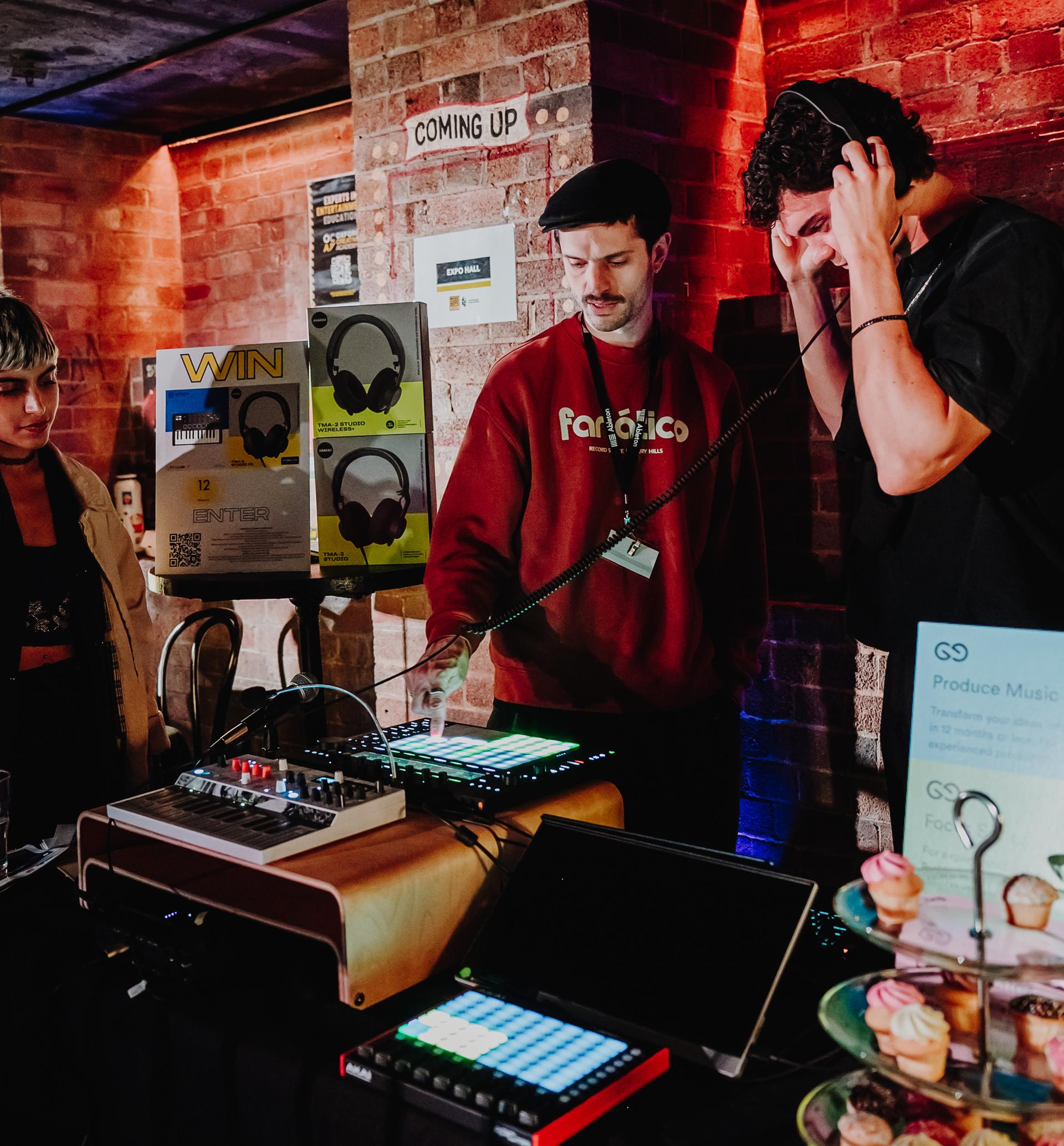 Three people interact with music equipment on a table in a dimly lit room. One wears headphones, and another operates controls on a device.