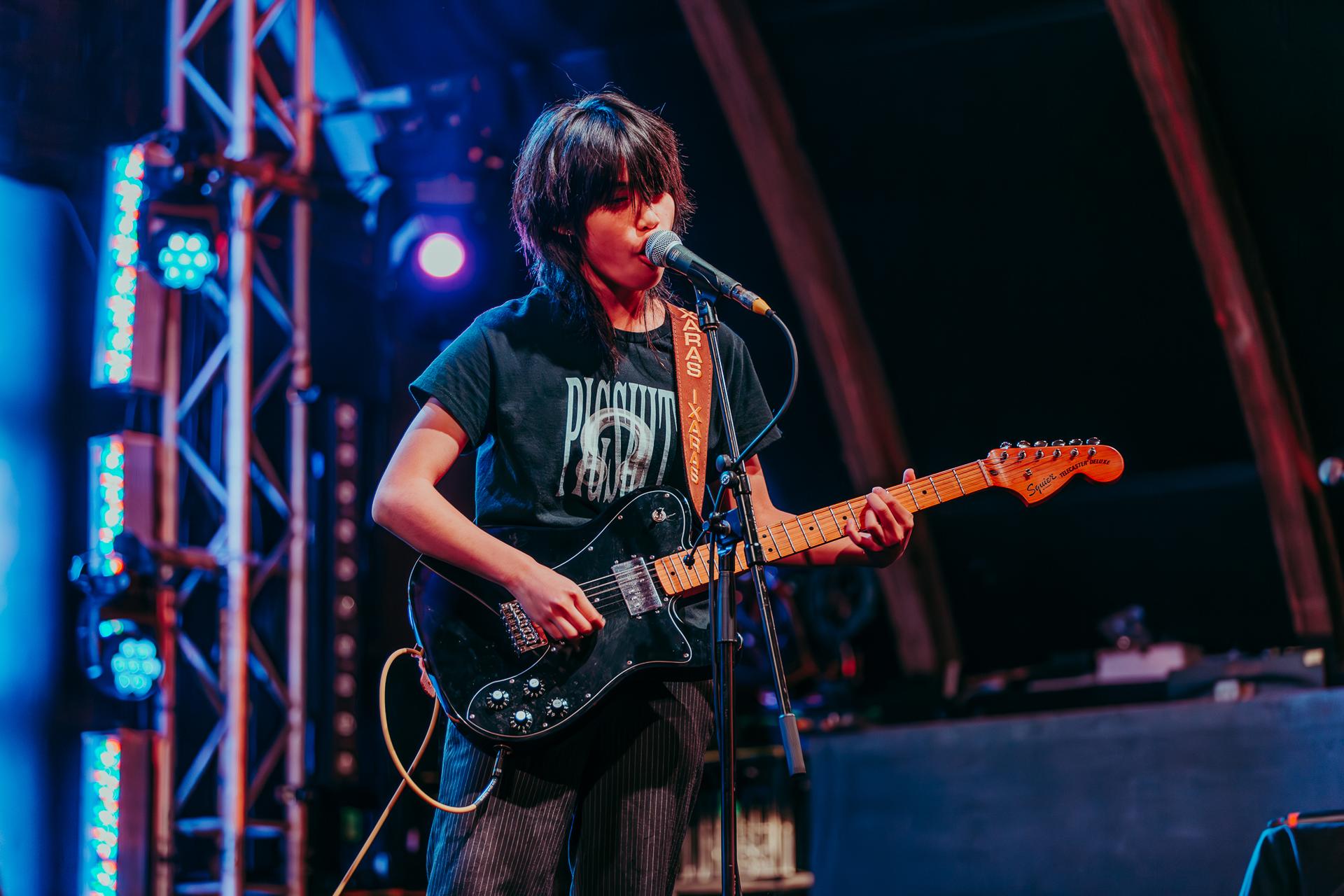 Musician on stage playing an electric guitar and singing into a microphone, with colorful stage lights in the background.
