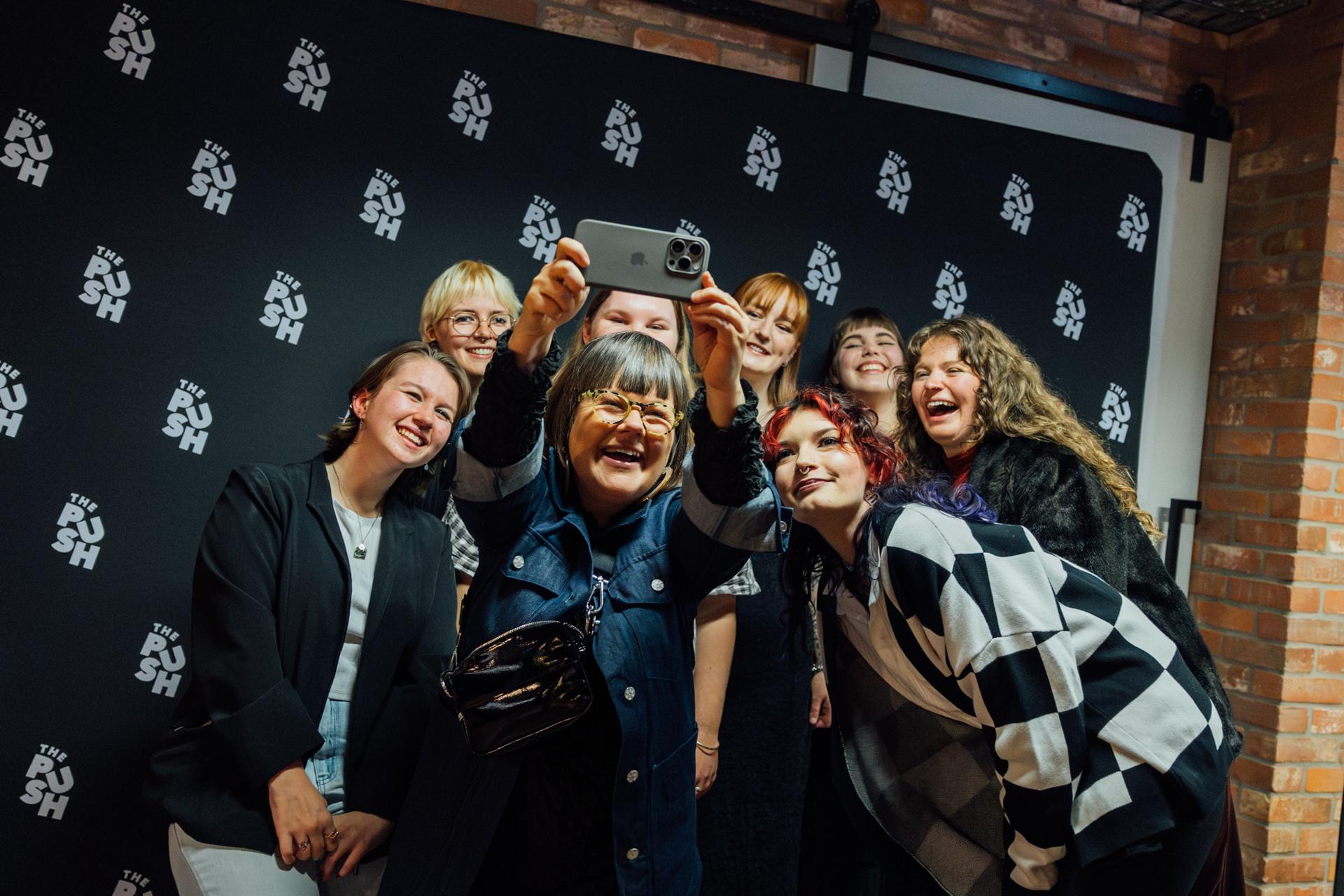 A group of people smiling and taking a selfie in front of a black wall with repeated "PUSH" logos.