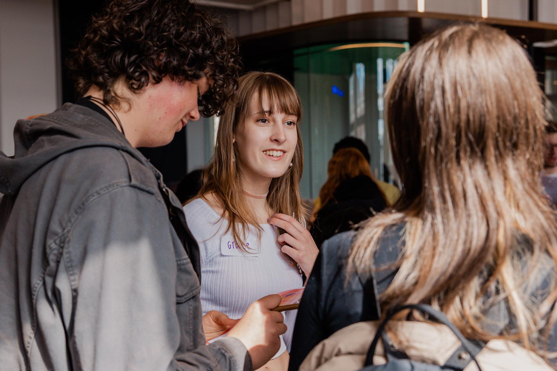 Three people are talking and smiling in a brightly lit indoor setting. One is holding a phone.